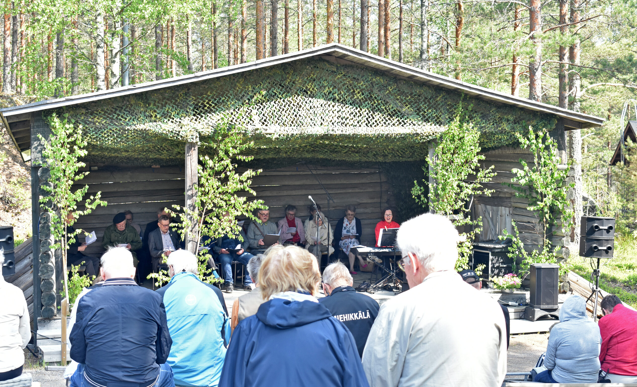 Yhteislaulutilaisuus menossa Salpalinja-museolla, näyttämöllä esiintyjiä ja soittajia, katsomossa yleisöä.